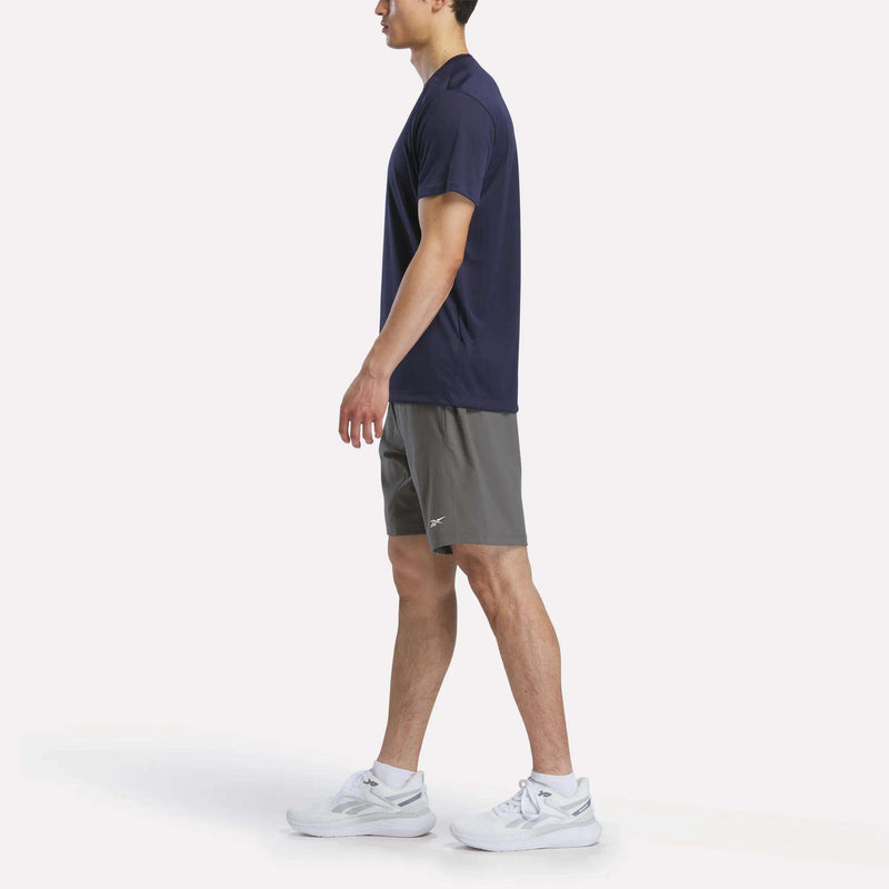 A man in profile wears a navy blue T-shirt, Reebok Men's Woven Shorts designed for workouts, and white sneakers against a plain white background.