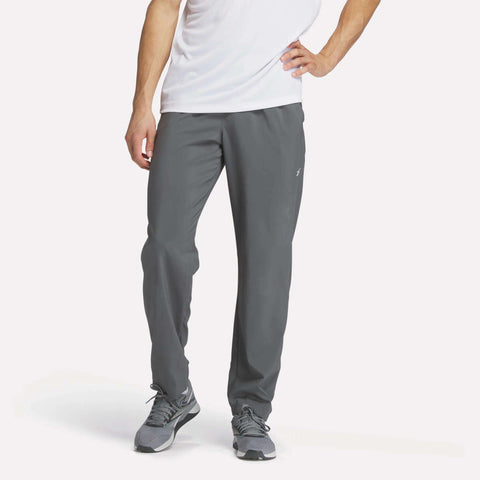 A person in a white T-shirt, gray sneakers, and Reebok Men's Train Woven Unlined Pants stands against a plain light background, with only the lower half of their face and body visible.