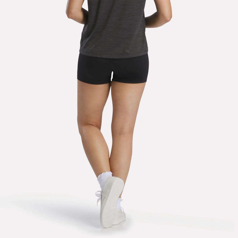 A person is shown from the back against a plain white background, standing with legs crossed and wearing a dark t-shirt, white socks, and white sneakers. They are also wearing black Reebok Women's High-Rise Mini Biker Shorts 3", which are designed with sweat-wicking fabric.