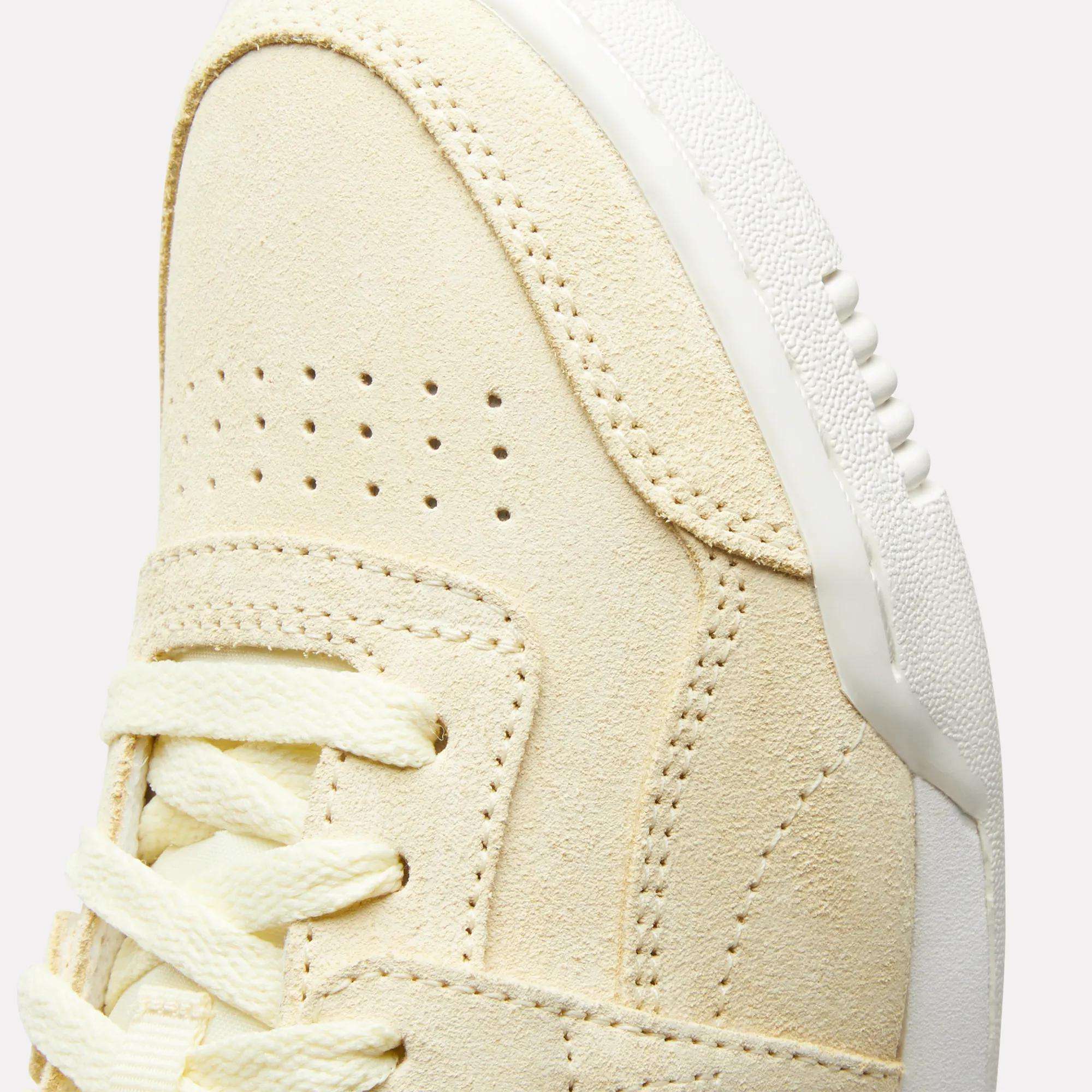 Close-up of the upper front of the Reebok Women's Workout Plus Shoes, featuring beige suede, double perforations, cream laces, and a white textured rubber sole—a classic '80s-inspired fitness style.