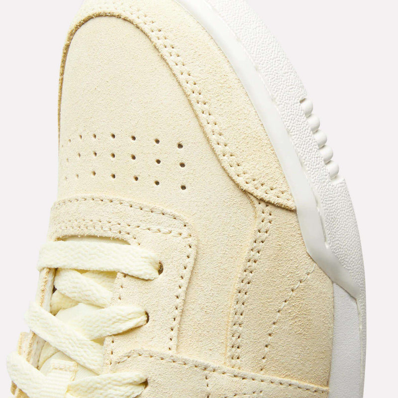 Close-up of the upper front of the Reebok Women's Workout Plus Shoes, featuring beige suede, double perforations, cream laces, and a white textured rubber sole—a classic '80s-inspired fitness style.