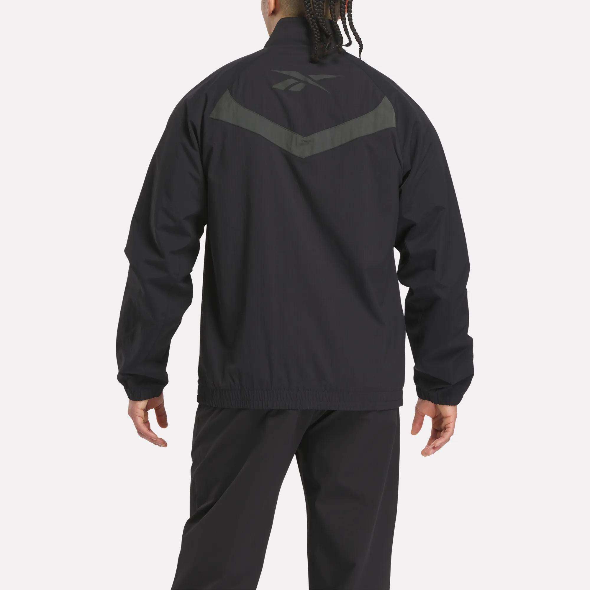 A person faces away, wearing a black Reebok Warm Up Track Jacket and pants. The jacket has a dark gray Reebok logo and chevron on the upper back. The background is plain and light-colored.