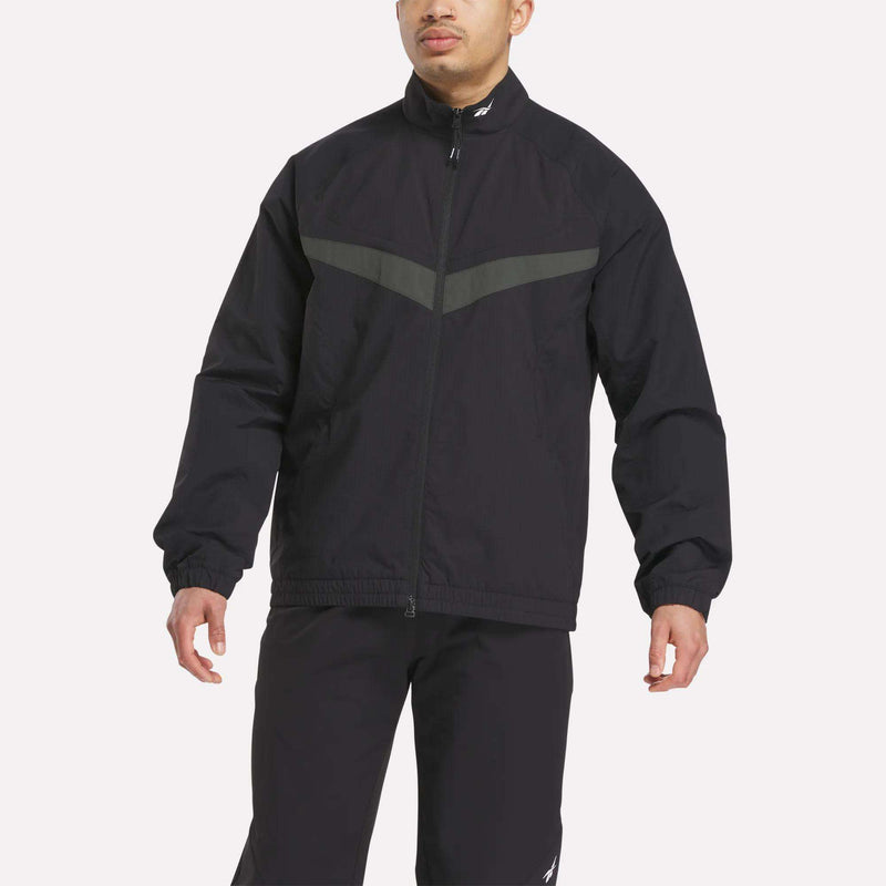 A person wearing the Reebok Warm Up Track Jacket in black with a gray chest stripe and matching black pants, standing against a plain light background.