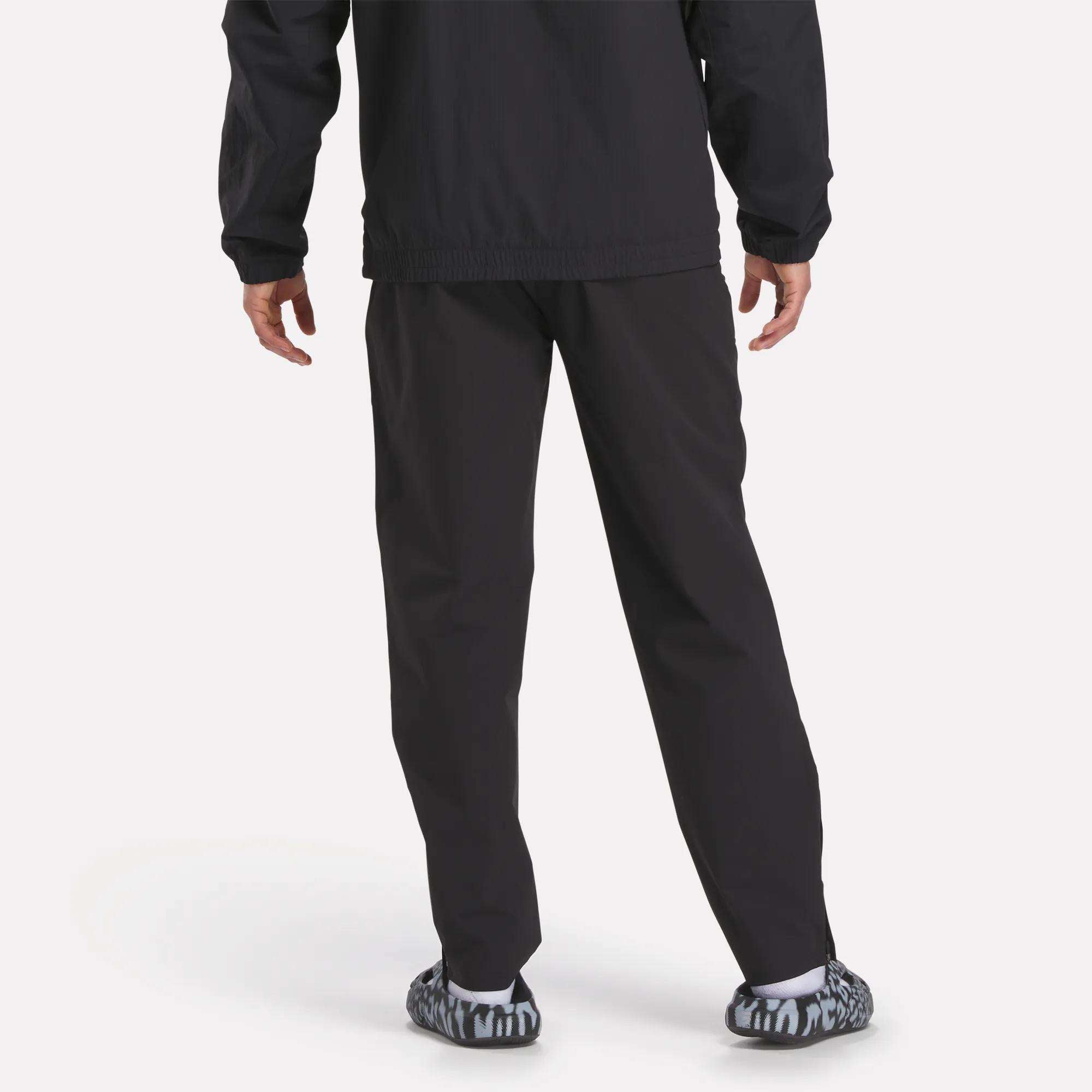 A person stands facing away in Reebok Track Pants, paired with a black jacket and black-and-white patterned slip-on shoes, against a plain light background.