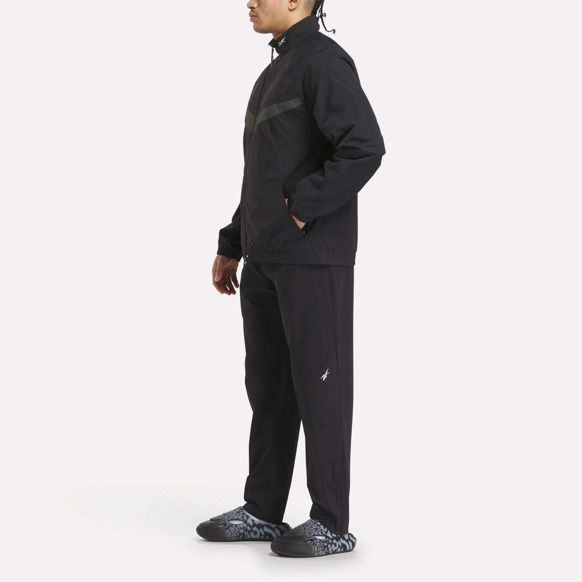 A person in profile wears Reebok Track Pants with a matching zipped jacket and patterned black and gray sneakers, standing against a plain white background.