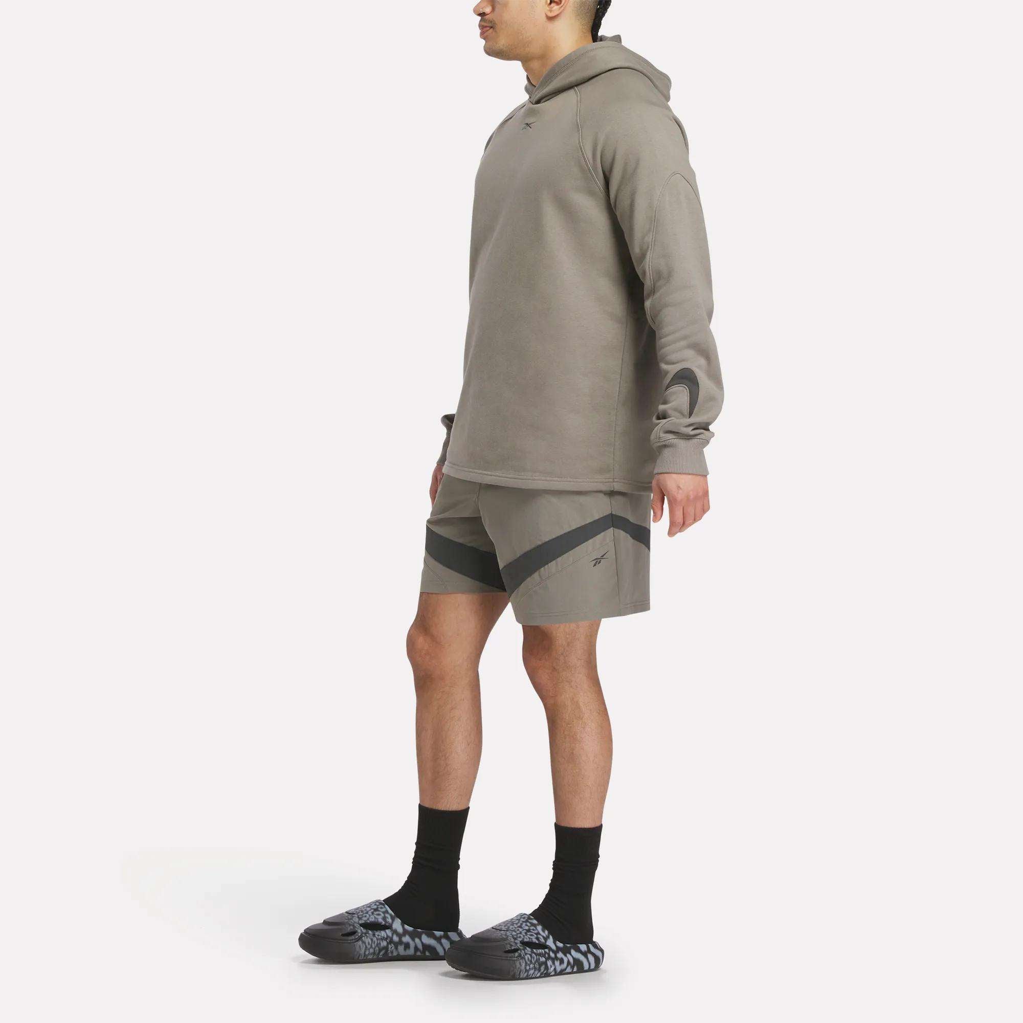 A person stands sideways against a plain white background wearing a taupe Reebok hoodie, Reebok Woven Basketball Shorts, black socks, and gray-patterned slip-on shoes.