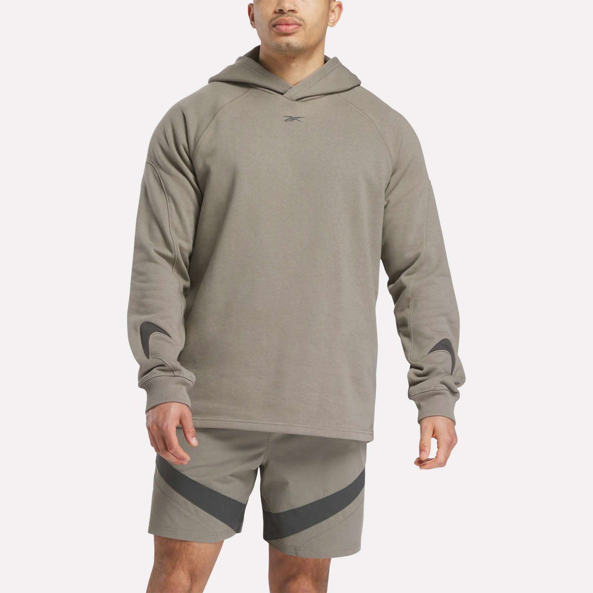 A man wears a light brown Reebok French Terry Basketball Hoodie with a small chest logo and matching shorts with dark diagonal stripes, standing against a plain white background.