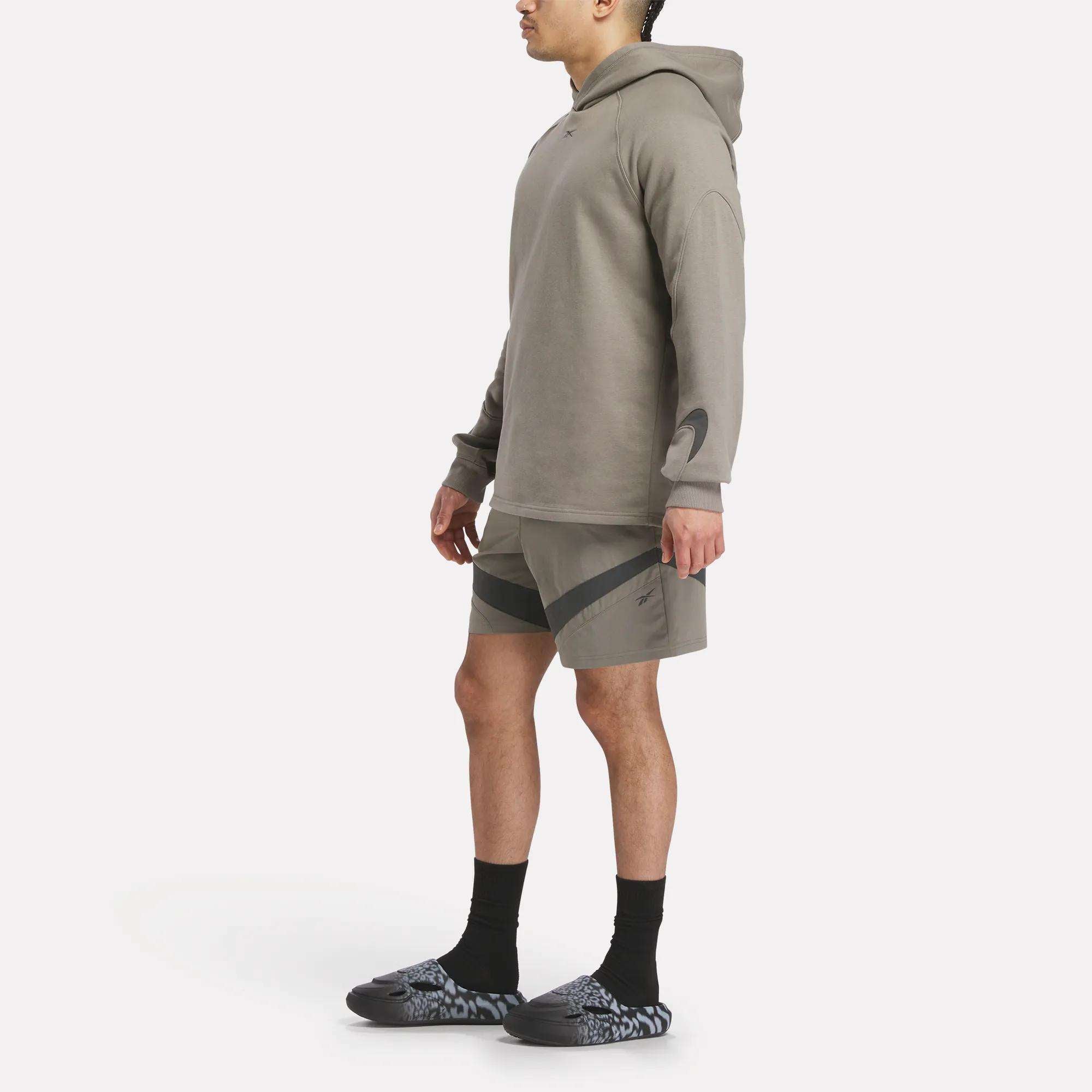 A person wearing a Reebok French Terry Basketball Hoodie in gray, matching shorts with black accents, black socks, and black-and-white slip-on shoes stands facing left against a plain white background.