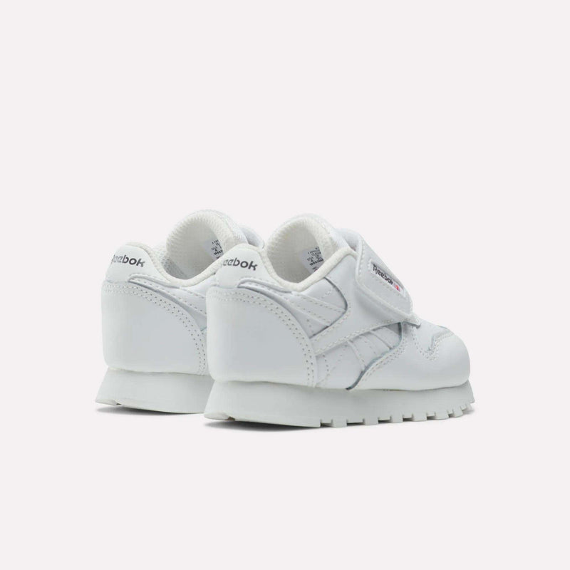 A pair of Kids' Classic Leather Shoes by Reebok, shown in white with Velcro straps, visible stitching, and rubber soles. Displayed from the back and side on a plain light background—ideal classic shoes for kids’ everyday wear.