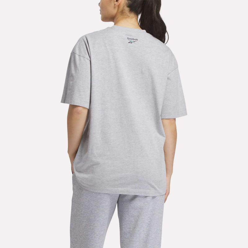 A woman in a loose-fitting Reebok Women's Lawn Club T-Shirt in light gray and matching pants stands facing away with her left hand in her pocket, dark hair tied in a ponytail.