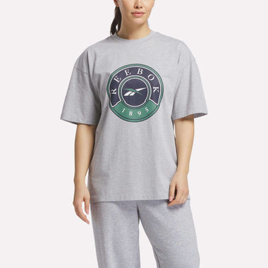 A woman wears the Women's Lawn Club T-Shirt by Reebok—a loose, light gray tee with a large circular Reebok logo and 1895 on the front, crafted from soft cotton knit for all-day comfort.