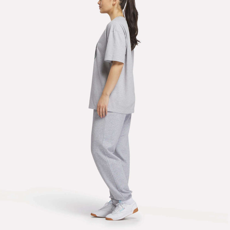 A person in profile wears a loose-fitting Reebok Women's Lawn Club T-Shirt, gray sweatpants, and white sneakers against a plain white background.