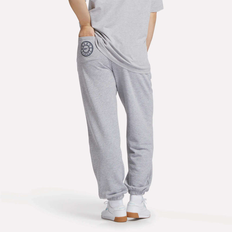 A person is seen from the back wearing Reebok Women's Lawn Club Jogger Pants in light gray, featuring an emblem on the pocket, paired with a matching gray tee and white sneakers, with hands tucked into the joggers' pockets.