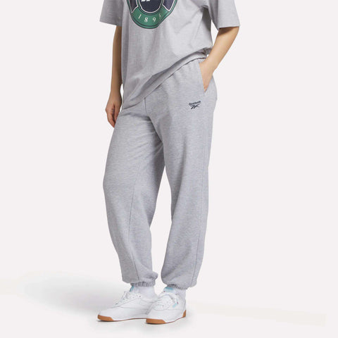 A person wearing a grey graphic t-shirt, Women's Lawn Club Jogger Pants by Reebok in light grey with a logo on the left thigh, and white sneakers, stands with hands in pockets against a plain background.