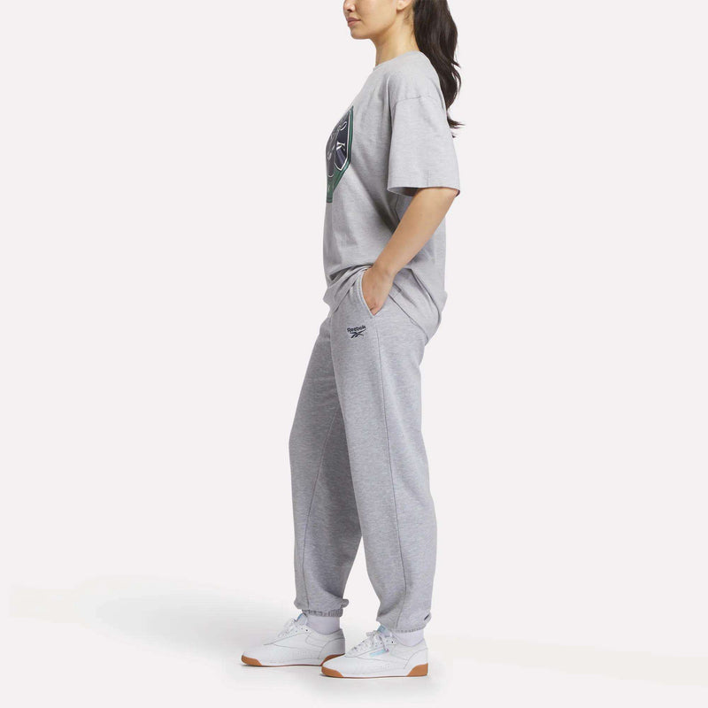 A woman stands in a relaxed pose wearing Reebok Women's Lawn Club Jogger Pants in grey, paired with a matching T-shirt and white sneakers. Her hands are in her pockets as she faces slightly to the side against a plain background.