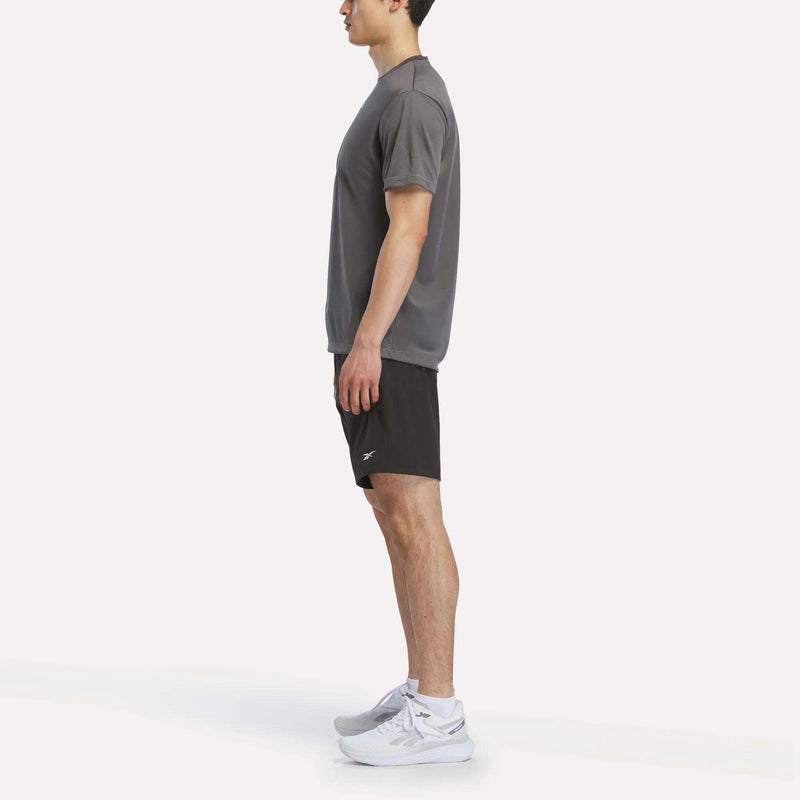 A man stands in profile against a white background, wearing a Reebok Men's Short Sleeve Tech Training T-Shirt in gray, black athletic shorts, and white running shoes.