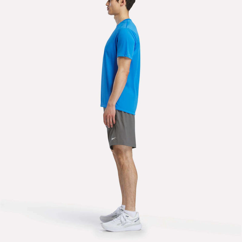A person stands in profile facing left, wearing a blue Reebok Men's Short Sleeve Tech Training T-Shirt, gray shorts, and white athletic shoes against a plain white background.