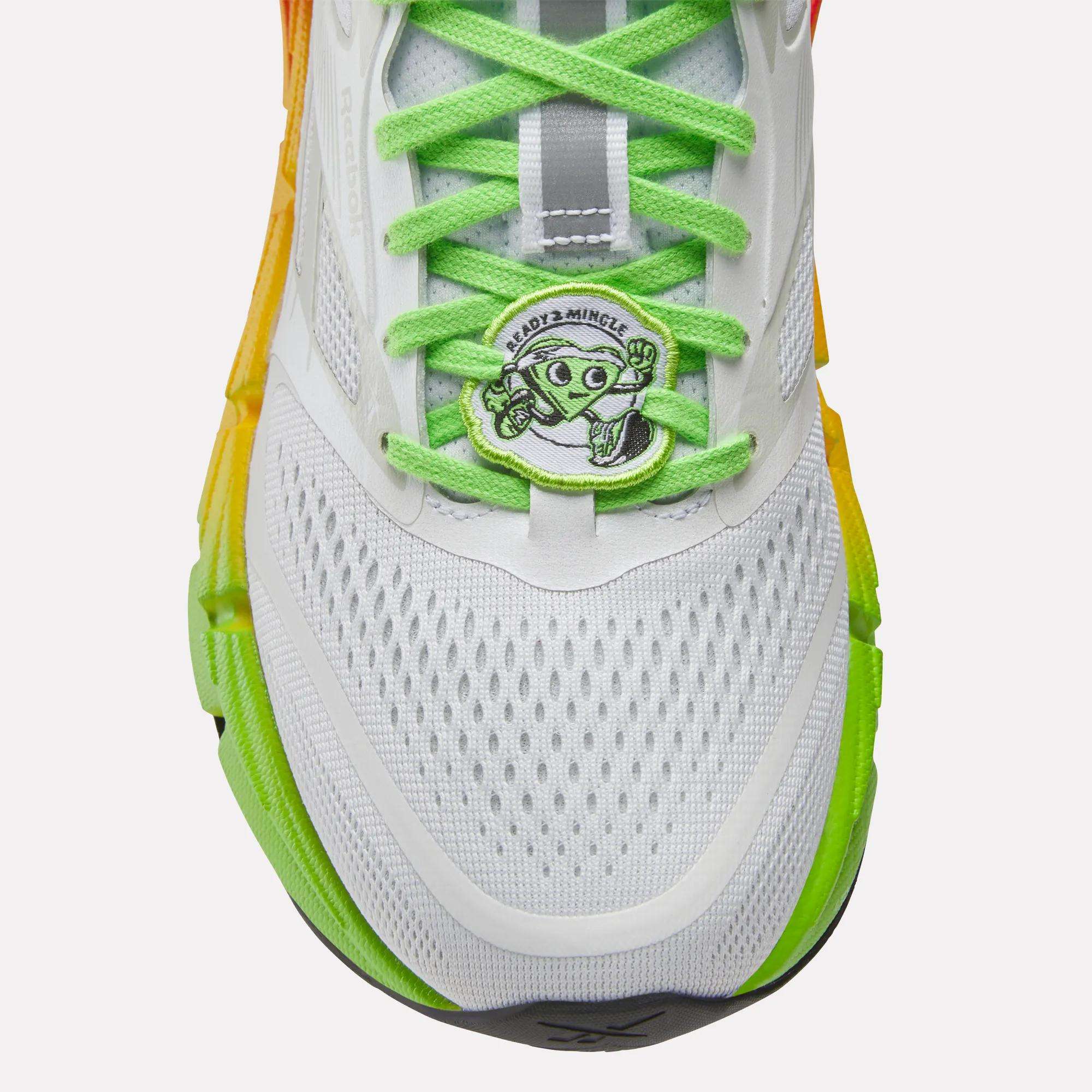 Close-up of Reebok Women's FloatZig 2 SoleMates Running Shoes in white with bright green laces, orange-yellow gradient sides, and a lightweight foam midsole. Features a cartoon alien peace-sign tag reading ANDO2+MINGLE.