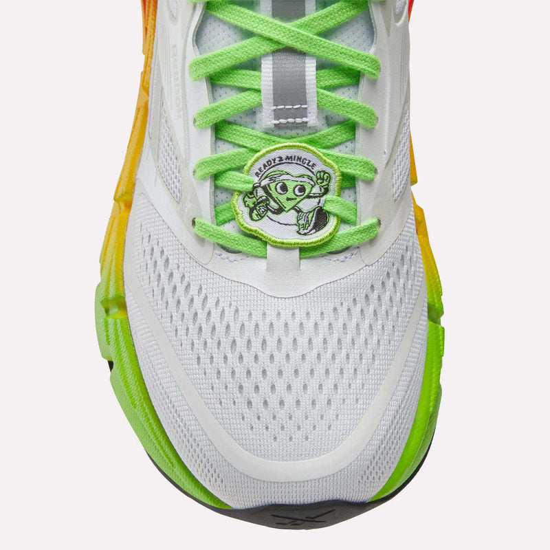 Close-up of Reebok Women's FloatZig 2 SoleMates Running Shoes in white with bright green laces, orange-yellow gradient sides, and a lightweight foam midsole. Features a cartoon alien peace-sign tag reading ANDO2+MINGLE.