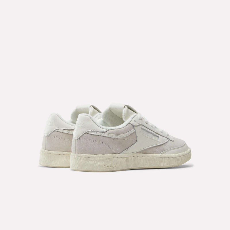 The Reebok x IDEA Club C 85 Shoes are displayed from a rear three-quarter angle, highlighting their off-white and light beige colorway inspired by the collaboration with IDEA bookstore. Crafted with a mix of suede and leather textures, these Reebok sneakers feature a cream rubber sole accented by subtle glow-in-the-dark details. Embossed Reebok branding is visible on the heel and side, while the shoes are set against a plain light gray background.