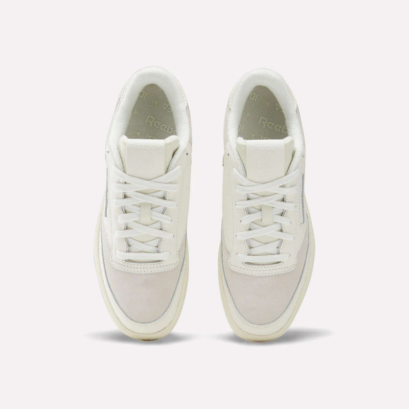 Top-down view of a pair of Reebok x IDEA Club C 85 Shoes, featuring cream-colored laces, suede toe boxes, and signature Reebok branding on the tongue and insole. Set against a plain, light grey background, this design blends effortlessly with the nostalgic vibe you’d expect to find among vintage books at the IDEA bookstore.