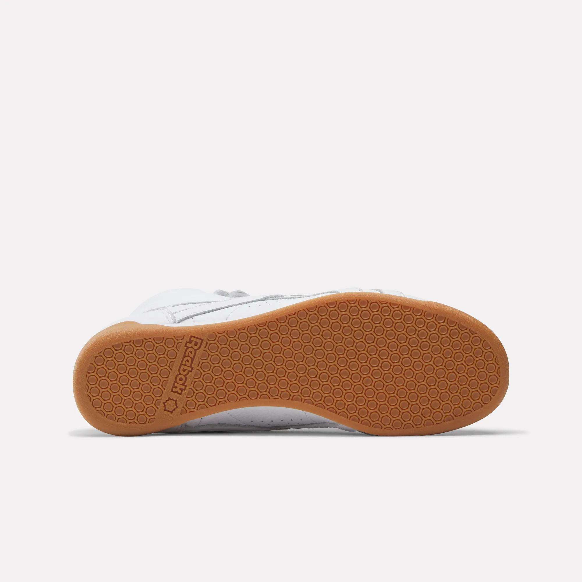 The Reebok Women's Freestyle Hi Shoes are shown from the bottom, showcasing a white upper, light brown gum sole with a hexagonal tread pattern, and the Reebok logo near the heel—a tribute to 80s fitness classics.