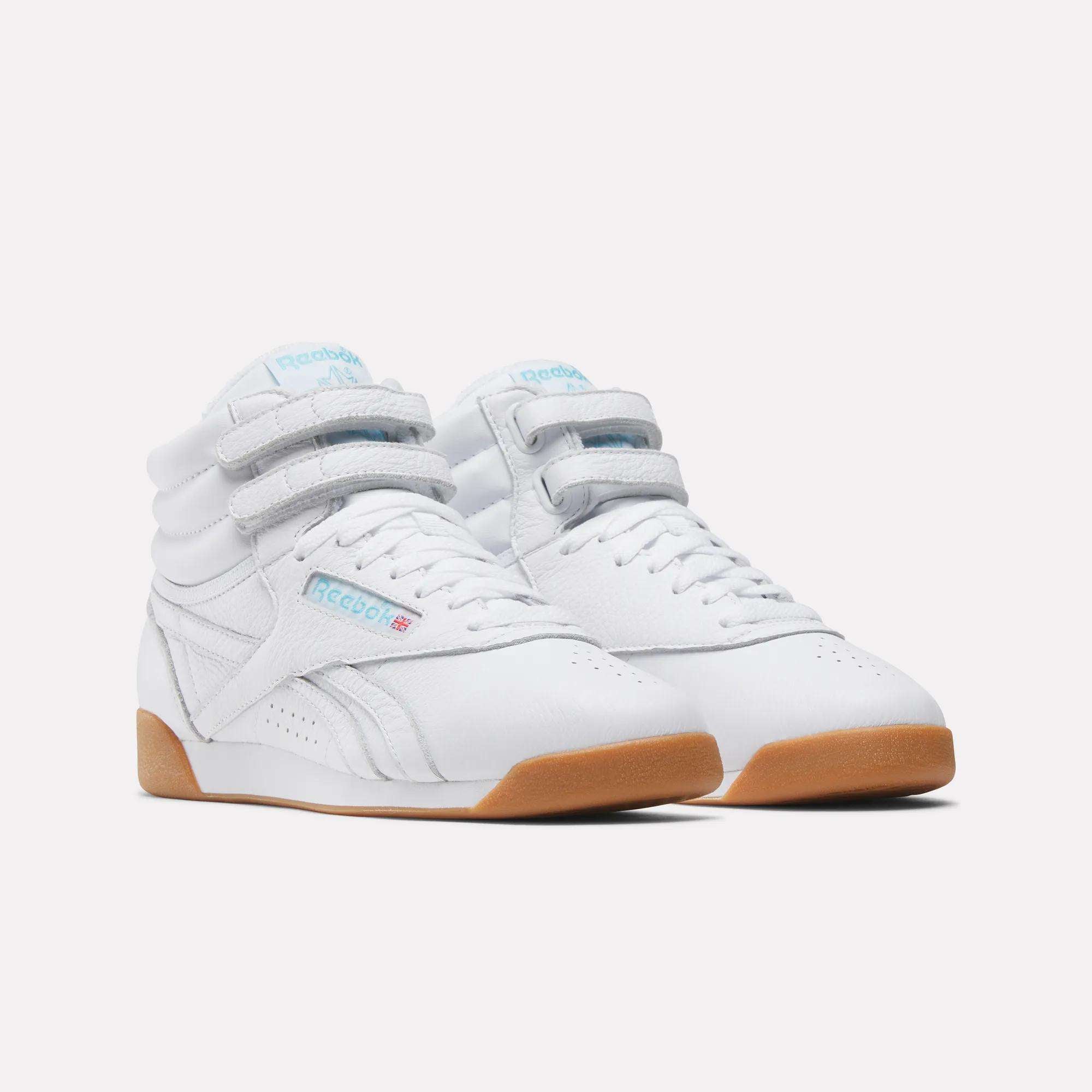 The Reebok Women's Freestyle Hi Shoes feature white high-top design, Velcro straps, light blue branding, and tan rubber soles—an iconic 80s fitness classic—shown on a white background.
