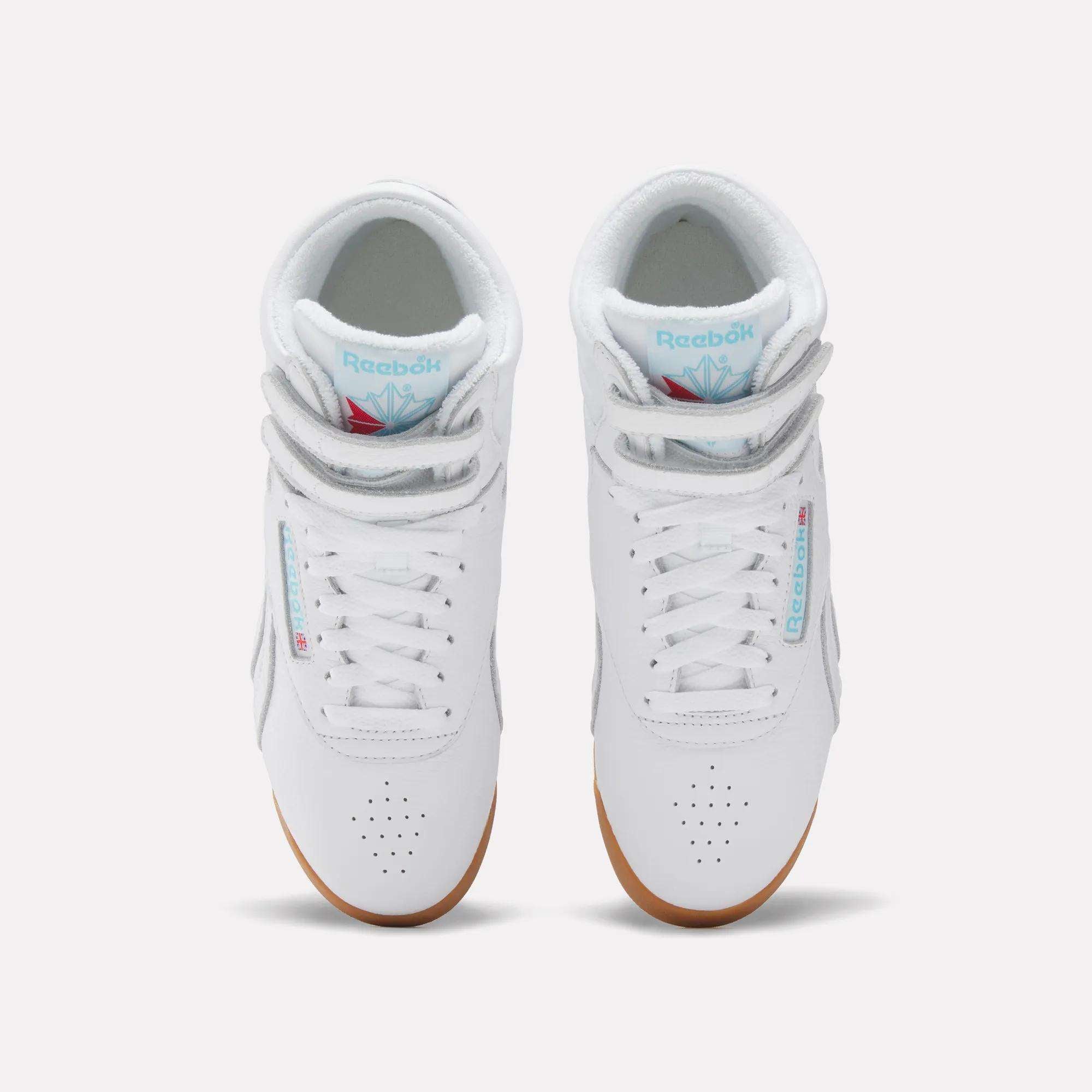 A pair of Women's Freestyle Hi Shoes by Reebok in white with gum soles, white laces, and blue Reebok logos on the tongue—an 80s fitness classic seen from above on a plain light background.