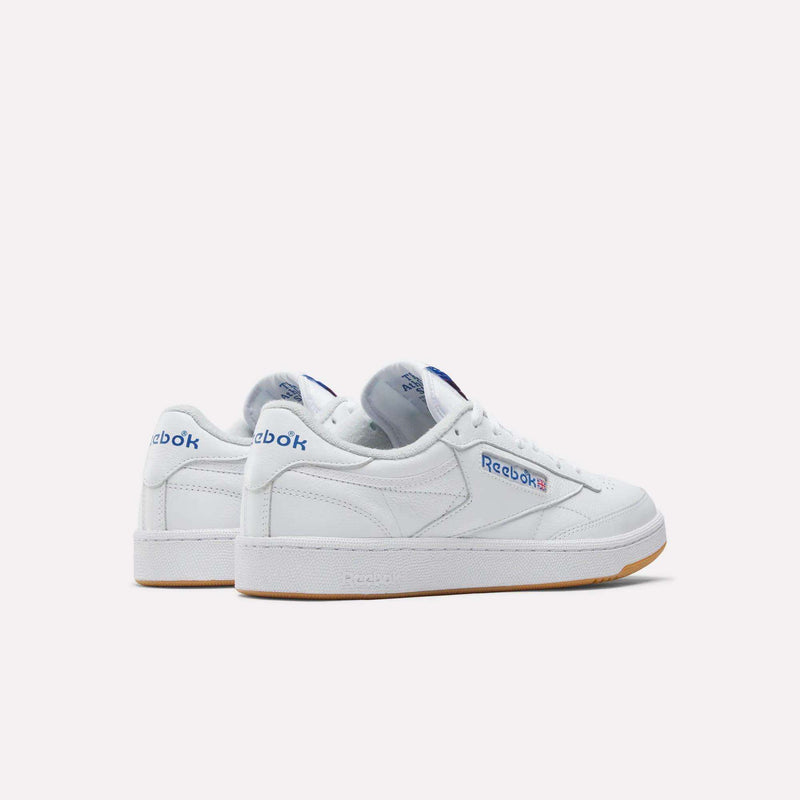 The Men's Club C 85 Shoes by Reebok are white leather low-tops with blue and red branding, tennis-inspired design, and gum rubber soles, shown on a light background.