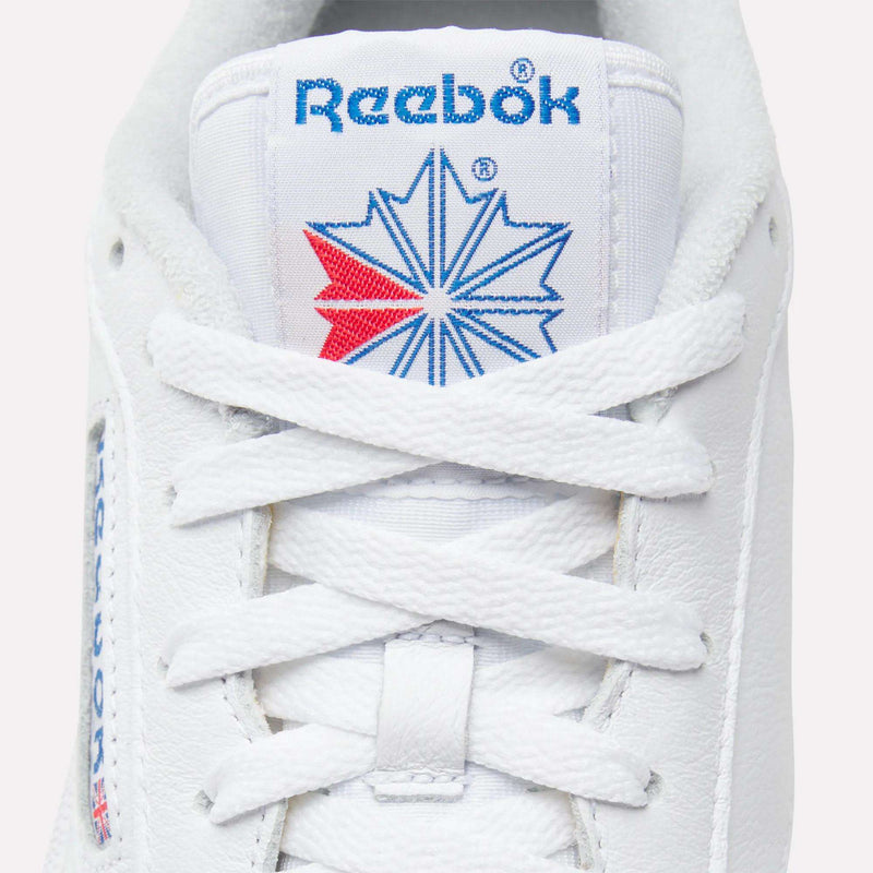 Close-up of Men’s Club C 85 Shoes by Reebok, featuring white leather, neat white laces, and the tongue displaying a blue Reebok logo with a red and blue starburst graphic.