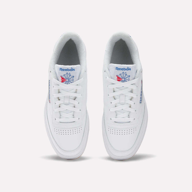 The Reebok Men's Club C 85 Shoes feature a white leather upper, blue Reebok branding, a red and blue star logo on the tongue, white laces, and perforated toe details for a classic tennis-inspired look.