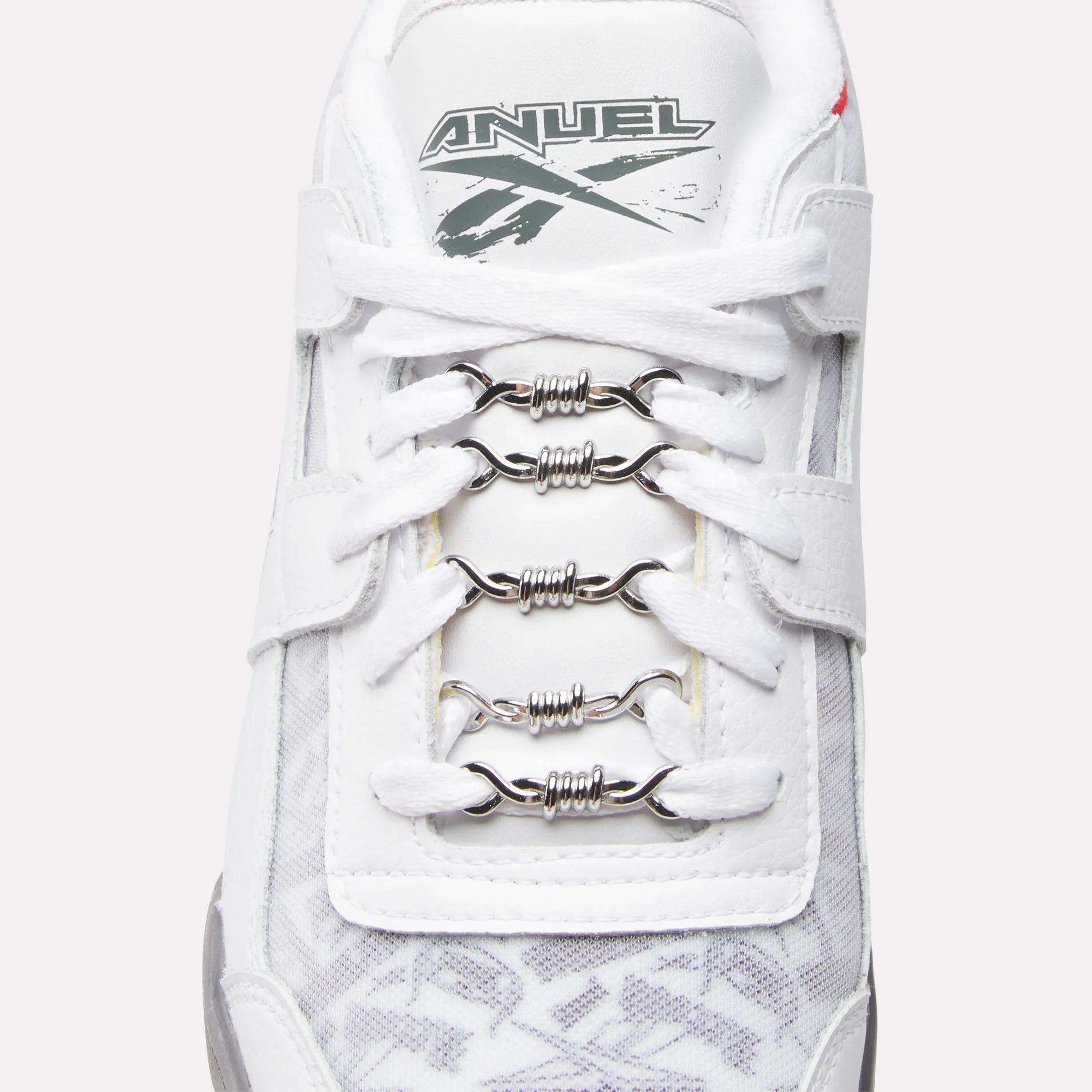 A close-up of a white Reebok Women's Zignition Running Shoe with metallic barbed wire-shaped lace accessories and "ANUEL" plus a gray graphic on the tongue, fusing bold style with cushioned running comfort.