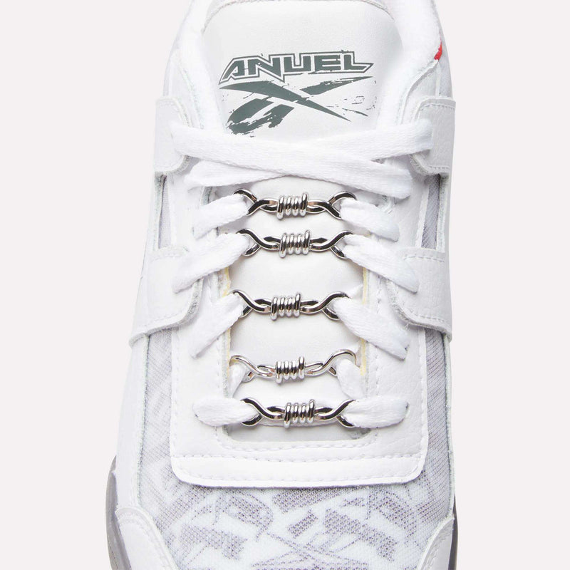A close-up of a white Reebok Women's Zignition Running Shoe with metallic barbed wire-shaped lace accessories and "ANUEL" plus a gray graphic on the tongue, fusing bold style with cushioned running comfort.