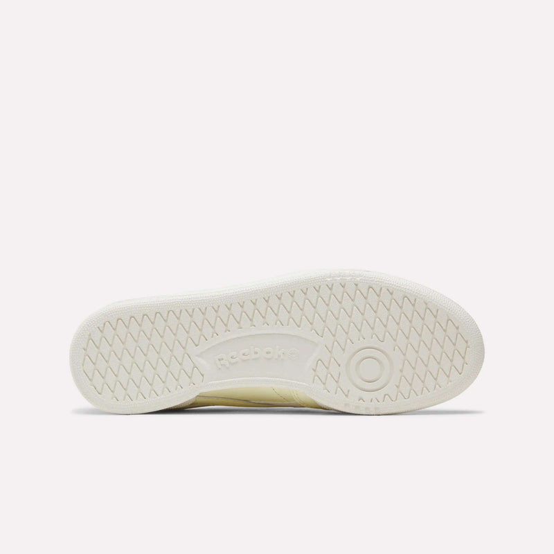 The image displays the bottom view of the Women's Club C Revenge Shoes by Reebok in a cream color, featuring a herringbone tread pattern and the Reebok logo at the center, perfectly capturing its vintage court style.