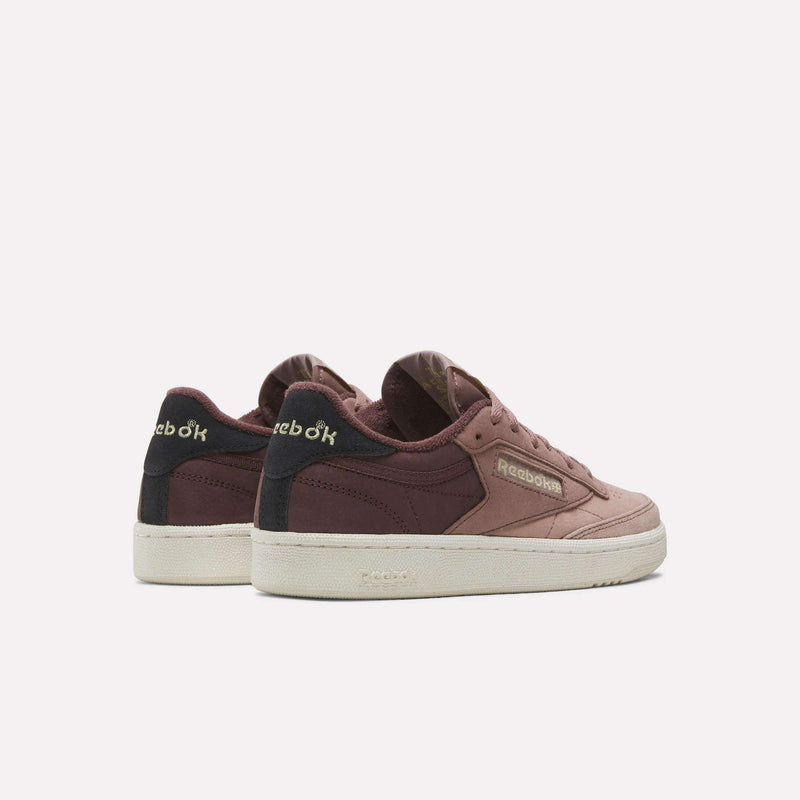 A pair of Reebok Women’s Club C 85 Shoes is displayed at a slight angle on a plain light background, featuring mauve and maroon nubuck leather uppers with cream soles. Designed as women’s tennis shoes, these sneakers showcase the yellow Reebok logo on both the side and heel.