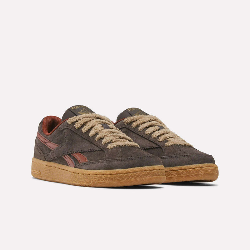 A pair of Reebok Women’s Club C Grounds 70 Shoes is displayed on a plain white background. Featuring a low-top suede construction in brown, these sneakers are detailed with beige fuzzy laces, retro-inspired accents in brown and red, and tan rubber soles. Drawing inspiration from the iconic Club C Grounds 70 silhouette, they offer a seamless blend of classic style and modern comfort.