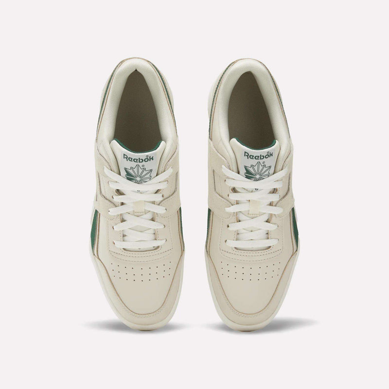 A top-view image of a pair of women’s Reebok Workout Plus Shoes in off-white leather with green details, displayed on a plain light background. The sneakers feature laces, perforated toe boxes, and tongue tags showcasing the Reebok logo and green leaf.