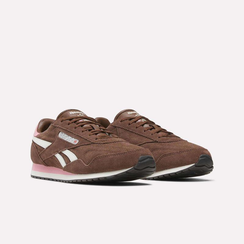 A pair of Reebok Women’s Classic AZ Shoes in brown, featuring white side stripes, pink accents on the heel and sole, white midsoles, and black outsoles are displayed on a plain white background.