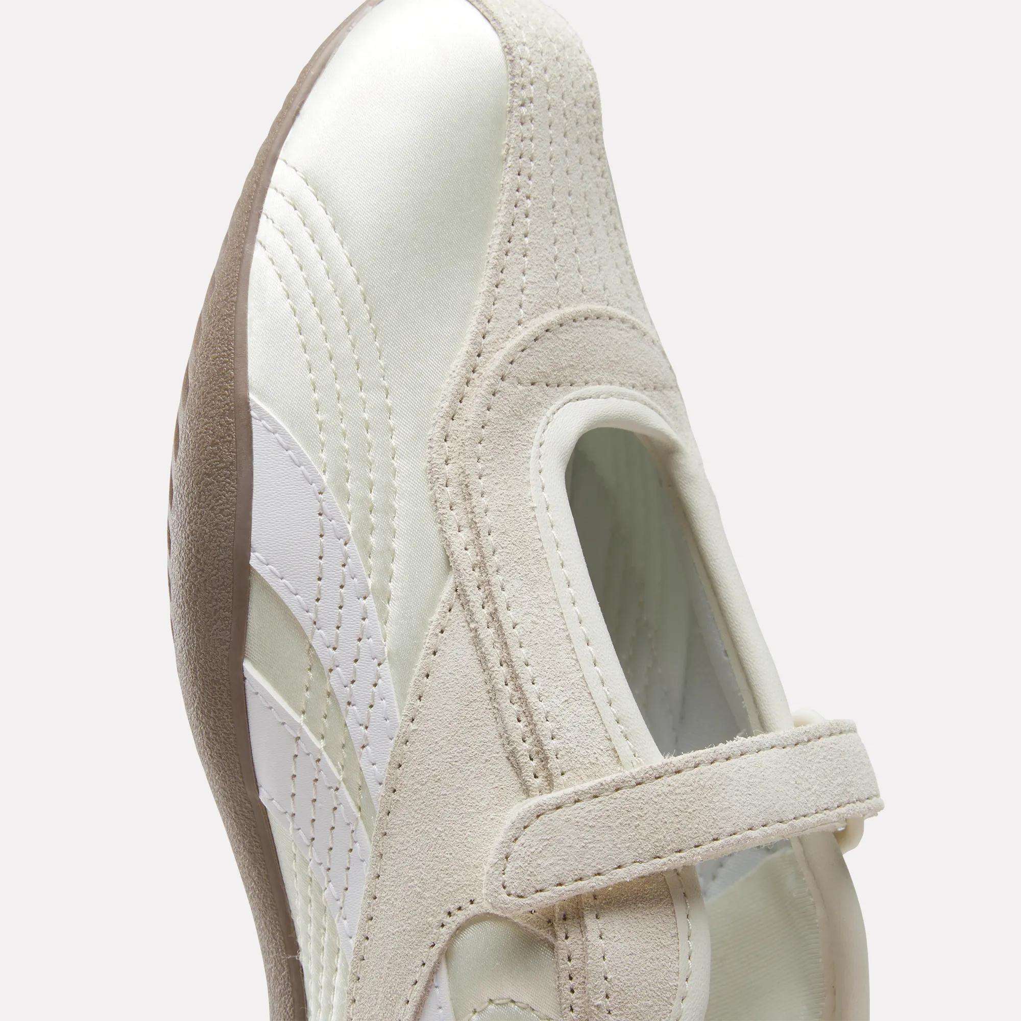 A close-up of the Reebok Women's Ultra Lo Mary Jane Shoes in white and beige, featuring a hook-and-loop strap, suede and leather accents, and a textured brown sole, shown against a light background for a modern, low-profile runner look.