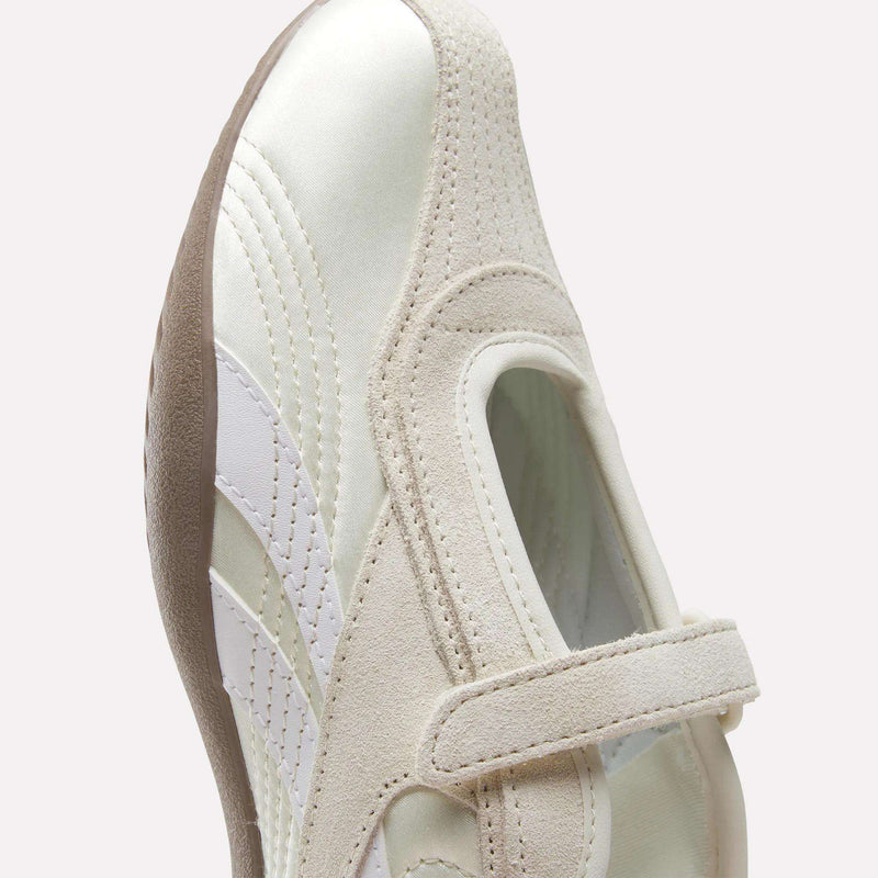 A close-up of the Reebok Women's Ultra Lo Mary Jane Shoes in white and beige, featuring a hook-and-loop strap, suede and leather accents, and a textured brown sole, shown against a light background for a modern, low-profile runner look.