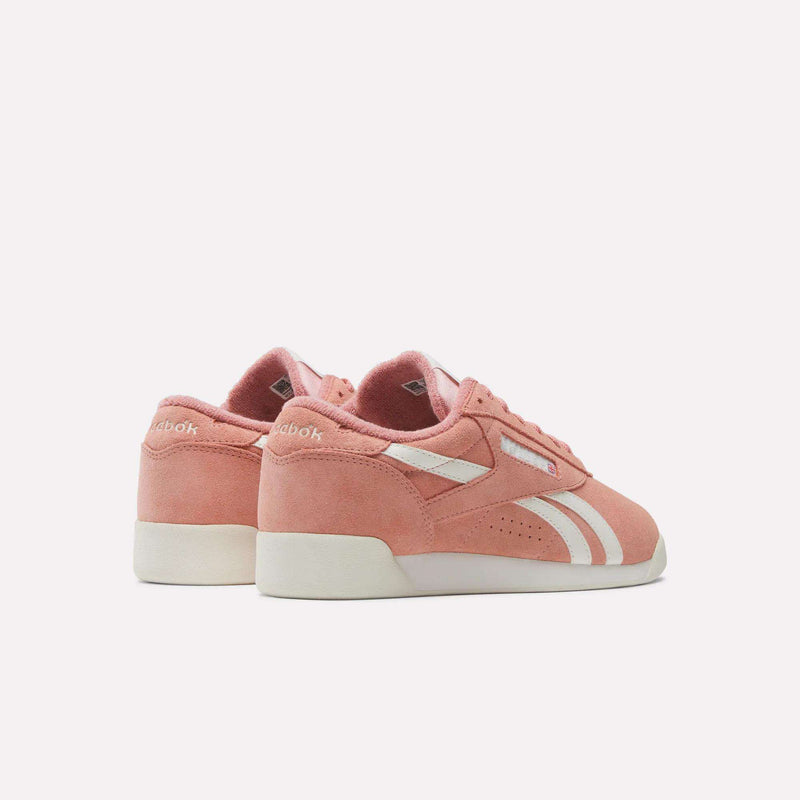 The image shows Reebok Women's Freestyle Lo Shoes in pink suede, featuring white soles and side stripes, displayed from the back and side angles against a plain white background.
