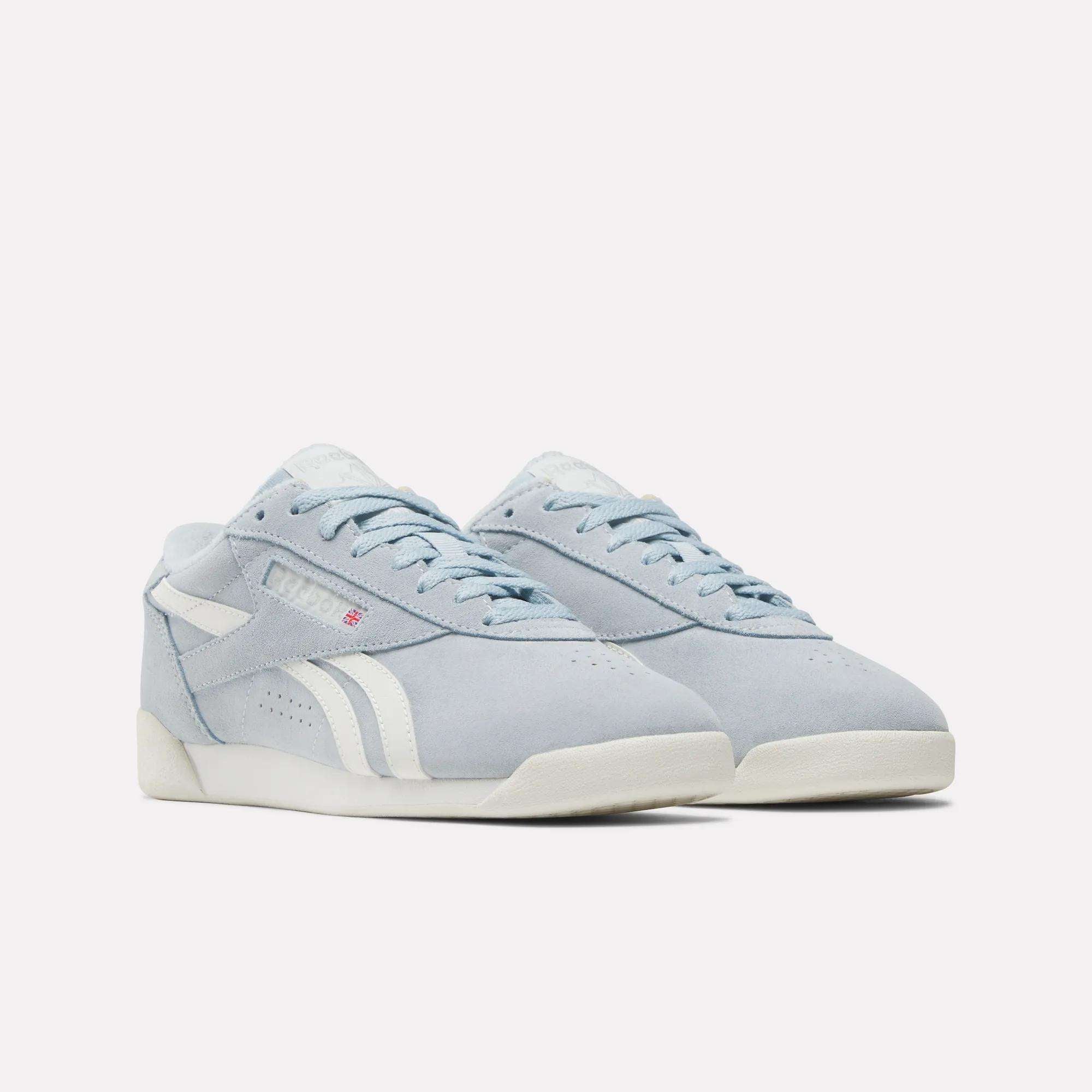 The Reebok Women's Freestyle Lo Shoes are light blue retro suede sneakers with white soles, white side stripes, laces, and a low-top design, shown on a plain white background.