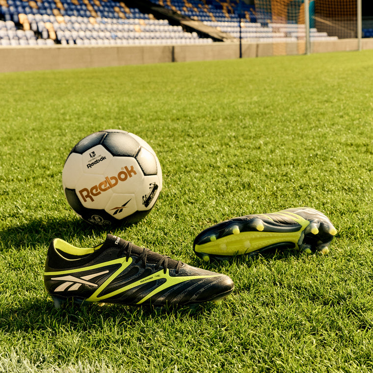 A black and white Reebok soccer ball and a pair of Sidewinder 26 Elite FG Cleats by Reebok, in black and yellow, rest on firm grass in an empty stadium with blue and yellow seats as a colorful backdrop.