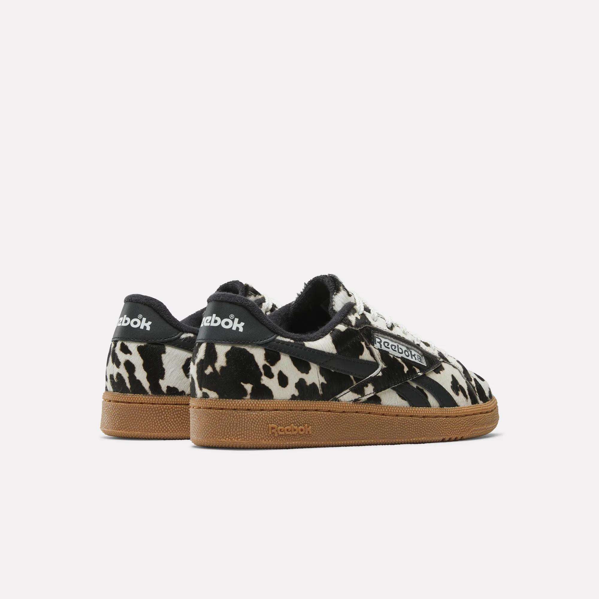The Reebok Women's Club C Grounds UK Shoes feature a bold black and white cow print, black heel tabs, white laces, and light brown rubber soles. Styled as women's sneakers, they are displayed against a plain white background.