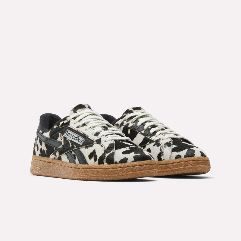The Reebok Women's Club C Grounds UK Shoes feature a bold black and white cow print and tan rubber soles, stylishly displayed on a white background.