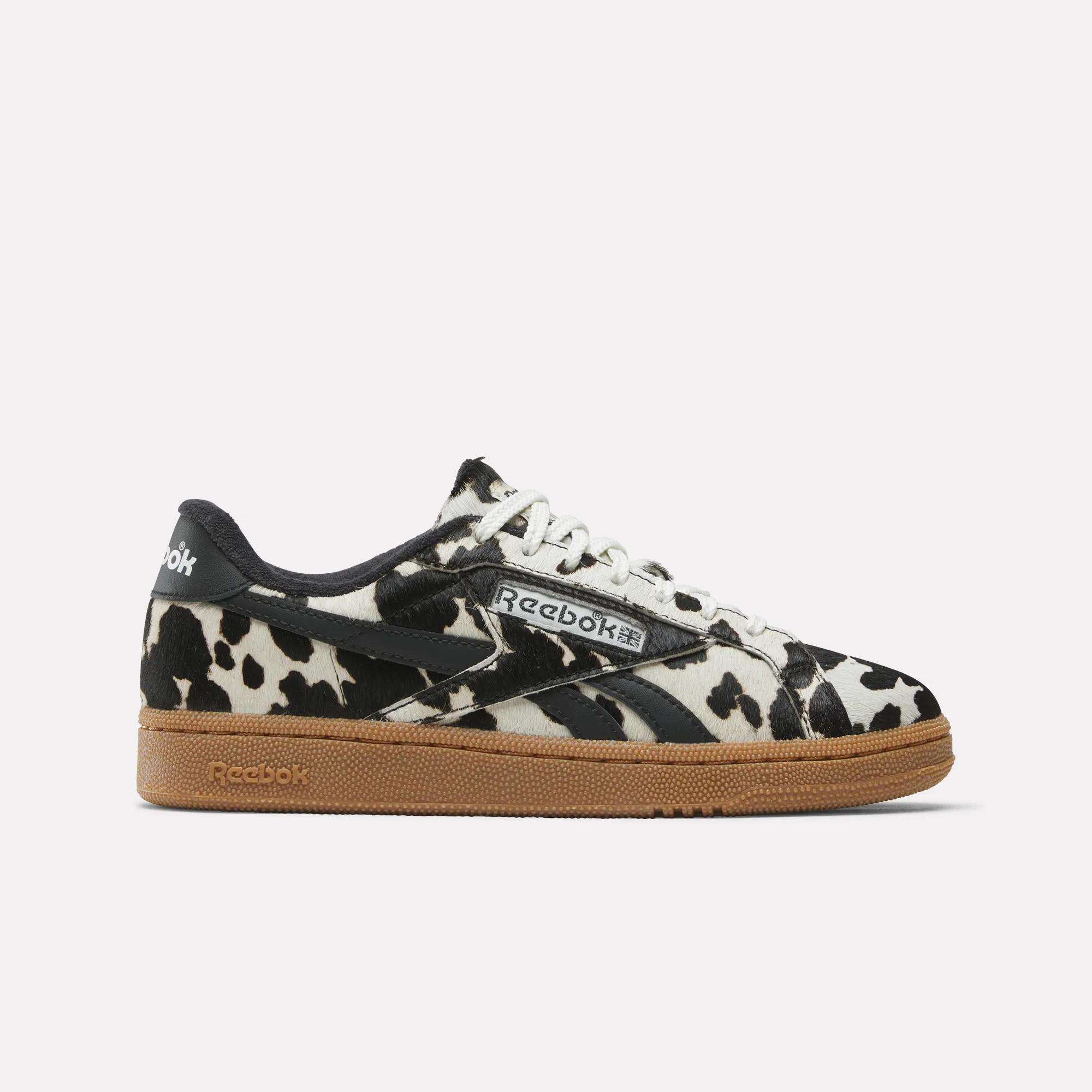 Women's Club C Grounds UK Shoes by Reebok feature a bold black and white cow print upper, white laces, black accents, and a brown gum sole, displayed on a plain white background.