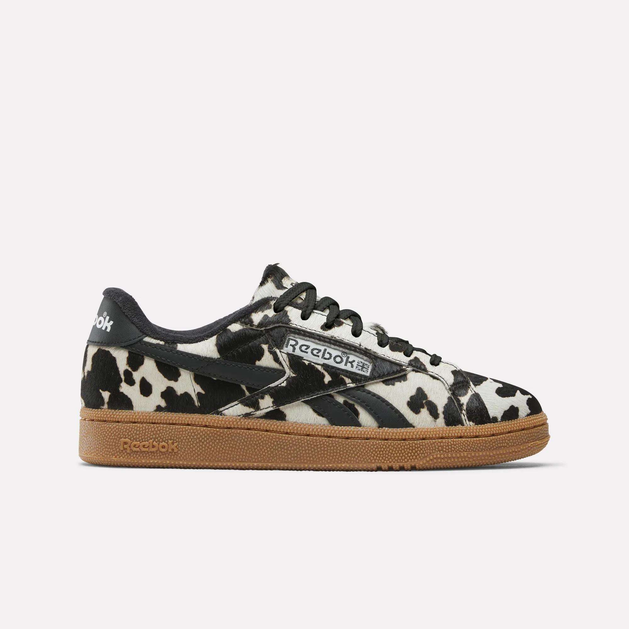 The Women's Club C Grounds UK Shoes by Reebok feature a bold cow print, black laces, black heel and tongue, plus a light brown gum sole—perfect for those who love unique Reebok sneakers.