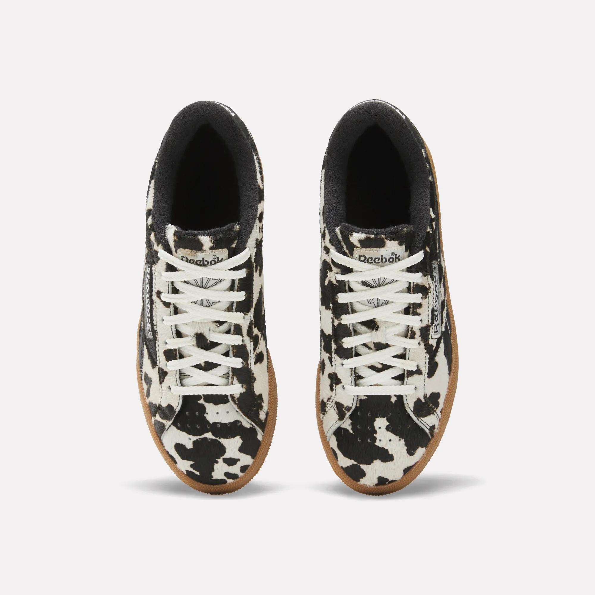 The Reebok Women's Club C Grounds UK Shoes feature a striking black and white cow print, white laces, gum soles, and a black inner lining. Shown from above on a plain light background.