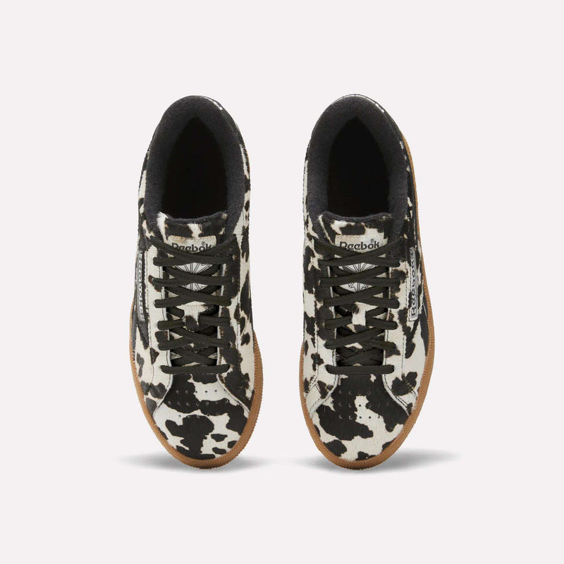 The Reebok Women's Club C Grounds UK Shoes feature a black and white cow print, black laces and lining, and brown outsoles, displayed from a top-down view on a plain background.