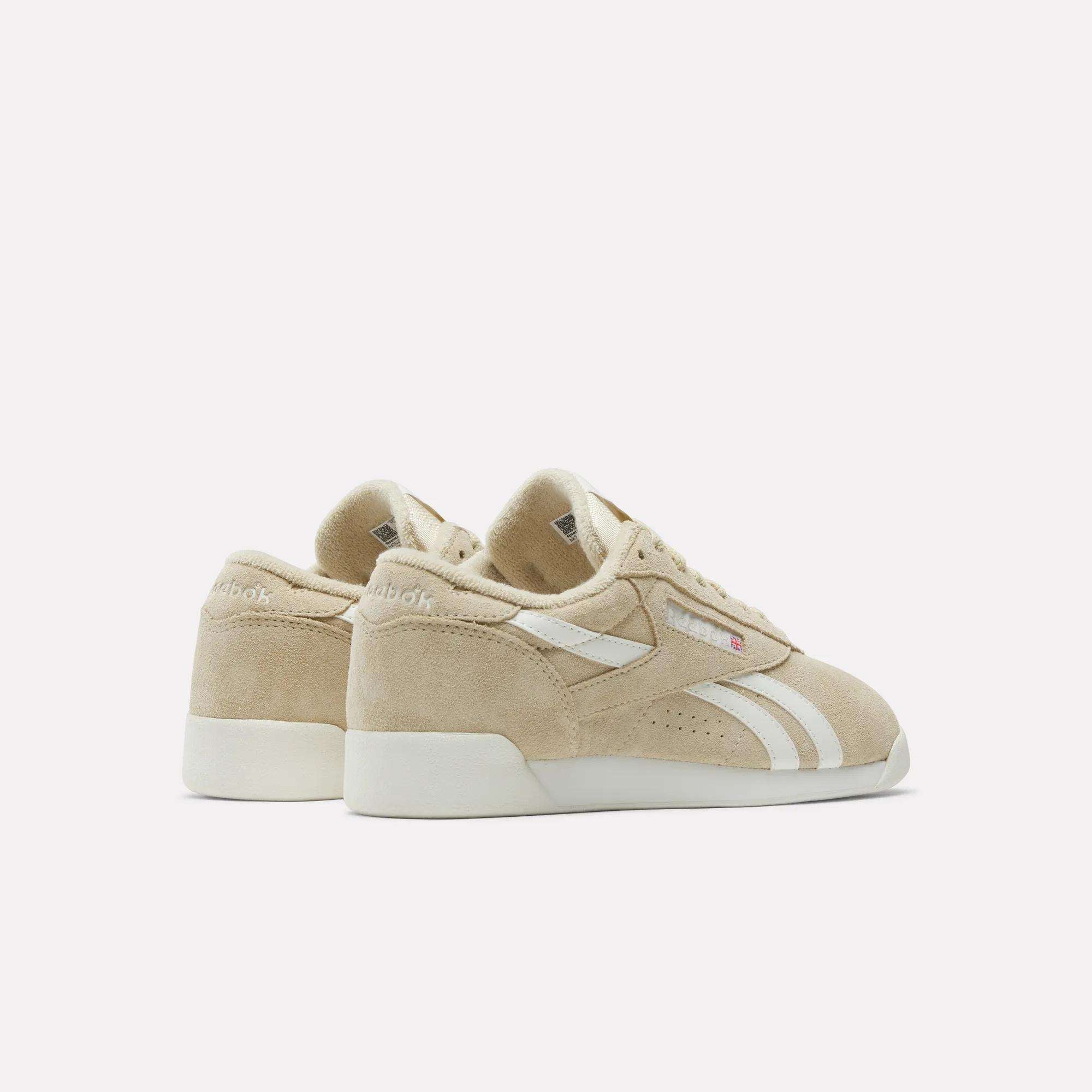 The Reebok Women's Freestyle Lo Shoes feature beige suede uppers, white side stripes, and rubber soles, shown from the back and side on a plain white background.